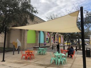 Shade covering and tables with seating with mural in background.