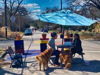Two people with dogs sitting at picnic table with umbrella, bench, and colorful barriers on residential street.