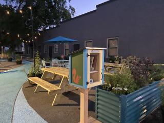 Little Free Library, planters, picnic tables, lights, umbrella, and ground mural.