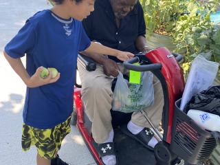 Young child helping holder adult on mobility scooter in community garden.